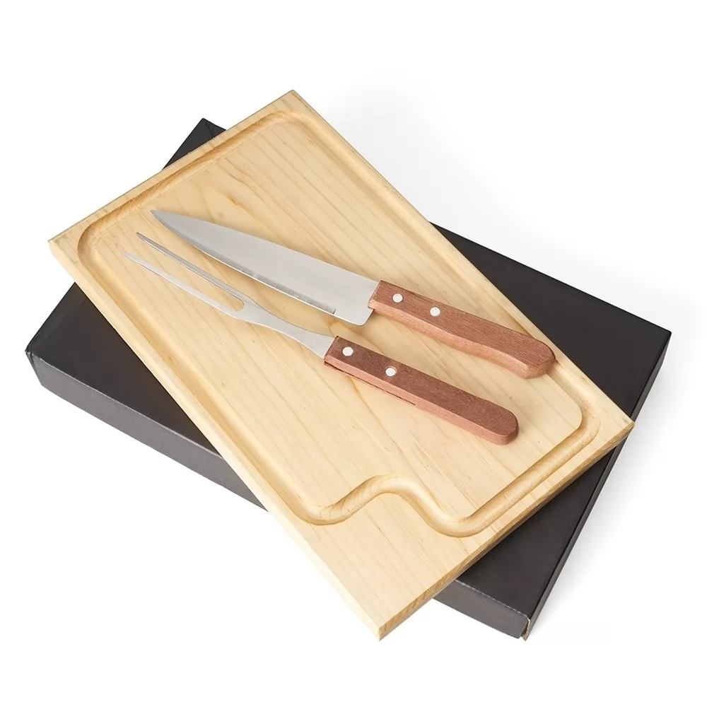 Kit churrasco com tábua de corte em madeira clara, faca e garfo sobre caixa preta em fundo branco
